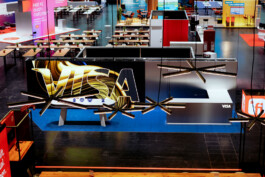 Exhibition hall with a large VISA booth at the center, viewed from an elevated angle. The booth features a bright VISA logo with golden wave patterns on a dark background. In the foreground, modern black ceiling lights with linear elements hang above. Additional booths and seating areas are visible in the background, including one with the text “Does AI have feelings?” keingarten