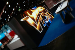 Tilted view of a trade fair setting featuring a large illuminated VISA logo on a black wall. Abstract exhibition booths and a ceiling with dynamic lighting are visible in the background. The logo is embedded in flowing orange-blue color gradients, evoking a sense of energy and motion. keingarten
